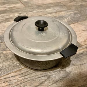 May include: A round, aluminum cooking pot with a lid. The pot has a black handle on top and two black side handles. The pot is sitting on a wooden surface.