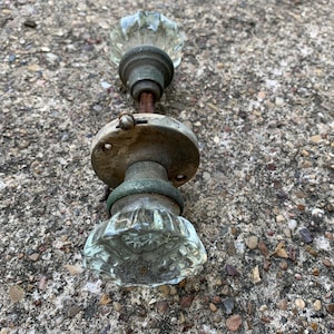 May include: An antique door knob set featuring clear, faceted glass knobs and aged metal hardware. The set includes two knobs connected by a threaded rod and a circular backplate, showcasing a vintage aesthetic. The metal has a weathered appearance.