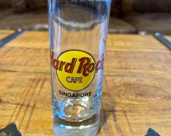 May include: A clear shot glass featuring the Hard Rock Cafe logo in red and yellow. The text "SINGAPORE" is printed below the logo. The glass is cylindrical and rests on a wooden surface.