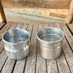 May include: Two small, silver-colored metal pots with handles. One pot has a rounded body with horizontal ridges, while the other is cylindrical. Both are sitting on a weathered wooden surface, likely a chair.