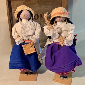 May include: Two handmade dolls dressed in traditional attire. One doll wears a blue skirt and carries a woven basket, while the other has a purple skirt and holds a bundle of straw. Both dolls have straw hats and are on wooden stands.