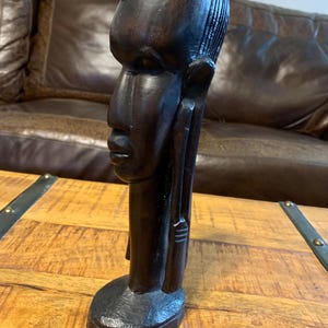 May include: A dark brown wooden sculpture of a person's head and upper body. The sculpture has a smooth finish and detailed facial features, including a prominent nose and defined lips. The figure has a long neck and a rounded base.