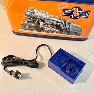 May include: An orange and blue Lionel Trains storage container with a black and white train graphic. A blue transformer with a dial and a black power cord are in the foreground.