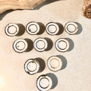 May include: A collection of nine circular cabinet knobs arranged in a triangle formation. Each knob features a smooth white center surrounded by a textured, metallic gold-toned rim with a rope-like pattern and visible signs of aged wear.