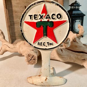 May include: A vintage Texaco sign, featuring a red star with a green "T" in the center, set against a white background. The sign has black lettering and a distressed, aged appearance. The sign is mounted on a white stand.