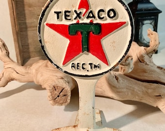 May include: A vintage Texaco sign, featuring a red star with a green "T" in the center, set against a white background. The sign has black lettering and a distressed, aged appearance. The sign is mounted on a white stand.