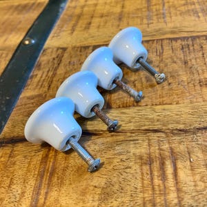 May include: Four white ceramic cabinet knobs with metal screws are arranged diagonally on a wooden surface. The knobs have a rounded, smooth shape and are slightly glossy. The screws are silver and show signs of age.