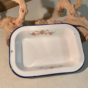 May include: A rectangular white enamelware dish with a blue rim and floral accents. The dish features a white interior with a blue border and decorative floral patterns in pink, yellow, and green. The dish is likely vintage.