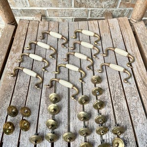 May include: A collection of vintage brass cabinet hardware, including handles with cream-colored center sections and matching round knobs. The hardware is arranged on a weathered wooden surface, showcasing a classic aesthetic.