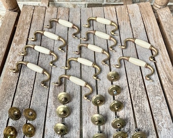 May include: A collection of nine curved drawer pulls featuring off-white ceramic centres with brass-toned metal ends, accompanied by twenty-two circular brass-toned metal cabinet knobs with central mounting points arranged in rows.