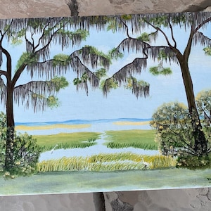 May include: An acrylic painting of a marsh scene with trees and Spanish moss. The painting features a blue sky, green marsh, and a white bird. The artist's signature and the year '86 are visible in the lower right corner.
