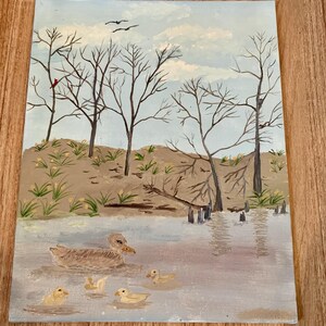 May include: An original painting of a serene waterscape featuring a mother duck and her ducklings swimming in a lake. Bare trees line the shore under a cloudy sky. The painting is done in muted tones of brown, beige, and blue.