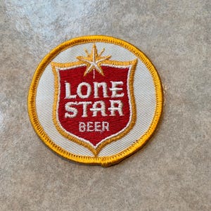 May include: Embroidered patch featuring the Lone Star Beer logo. The patch is circular, with a white background and a gold border. The logo is a red shield with the words "LONE STAR BEER" in white.
