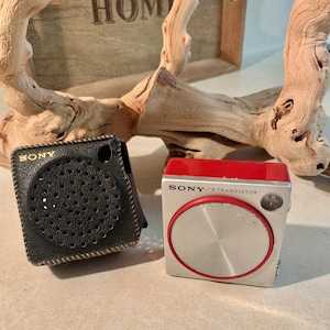 May include: Two vintage Sony transistor radios. One is black, square, and in a leather case. The other is silver with a red top and a red circle around the speaker. Both radios have the Sony logo.