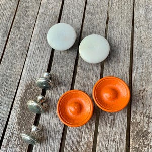 May include: An assortment of cabinet hardware, including four silver-toned knobs and two pairs of round, decorative pulls. The pulls are light green and orange. The hardware is arranged on a weathered wooden surface.