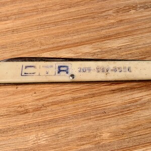 May include: A vintage folding knife with a cream-colored handle and a silver blade. The handle has the letters "CTA" and a phone number printed in blue. The knife is lying on a wooden surface.