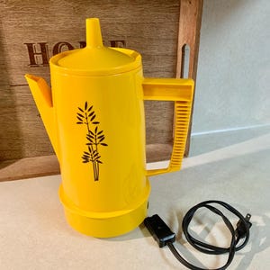 May include: A bright yellow electric coffee pot with a black floral design. The pot has a lid, a spout, and a handle. A black power cord is connected to the base. The pot is sitting on a white surface.