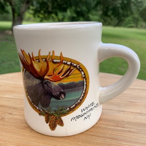 May include: White ceramic mug featuring a detailed illustration of a moose with large antlers set against a scenic background. The mug has a curved handle and the text "White Mountain, NH" is printed on the side. The mug is on a wooden surface.