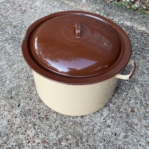 May include: A vintage enamelware pot with a brown lid and a cream-colored body. The pot has a handle on each side and a handle on the lid. The pot is sitting on a concrete surface.