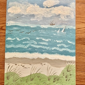 May include: An acrylic painting of a coastal scene. The artwork depicts a blue ocean with white-capped waves, a sandy beach, and a sky filled with fluffy white clouds. A sailboat and a fishing boat are visible on the water, with seagulls flying overhead.