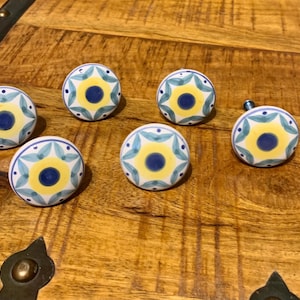 May include: Six ceramic cabinet knobs with a floral design. Each knob features a central blue circle surrounded by yellow and teal petal-like shapes on a white background. The knobs are arranged on a wooden surface.