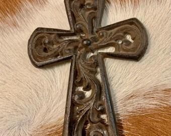 May include: An ornate, dark brown metal cross featuring detailed scrollwork. The cross is set against a backdrop of brown and white cowhide, enhancing its rustic appearance. A decorative religious item.