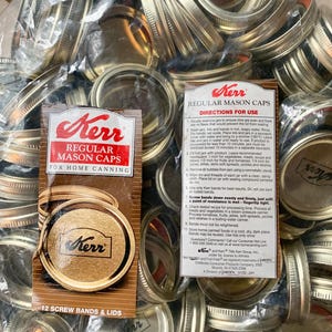May include: A collection of Kerr Regular Mason Caps for home canning. The image shows a clear plastic bag filled with metal screw bands and lids. A package of 12 screw bands and lids is visible, with instructions for use.