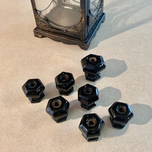 May include: A collection of eight glossy black hexagonal knobs with a threaded hole in the center. A decorative black and gold lantern is in the background. The knobs are likely for furniture or cabinet doors.