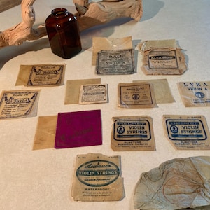 May include: An assortment of vintage violin string packets and a brown glass bottle. The packets vary in size and design, with text and graphics in various colors. Some packets are labeled "La Bella", "Helmet Violin Strings", and "Armour's Violin Strings".