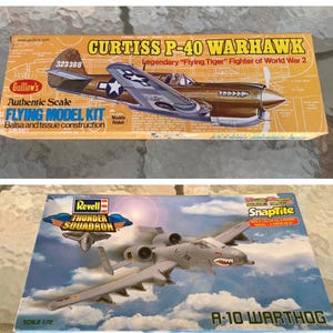 May include: Two model airplane kits are displayed. The top box is a Curtiss P-40 Warhawk, a Flying Model Kit with balsa and tissue construction. The bottom box is a Revell Thunder Squadron A-10 Warthog model kit. Both boxes feature detailed illustrations of the aircraft.