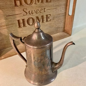 May include: A vintage silver-toned metal teapot with a curved spout and a handle. The teapot has a lid with a small knob on top. Behind the teapot is a wooden tray with the words "HOME Sweet HOME" engraved on it.