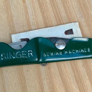 May include: A vintage green Singer sewing machine seam ripper with the brand name embossed on the side. The tool has a silver blade and a small paper insert. The words "SEWING MACHINES" are also visible.