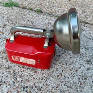 May include: A vintage red Big Beam Model 166 spotlight with a chrome handle and reflector. The spotlight has a rectangular base and is designed for portable use. The text "Big Beam Crystal Lake, IL 60014" is visible on the side.