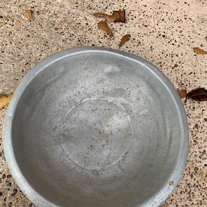 May include: A round, silver-colored metal bowl with a wide rim. The bowl has a slightly textured surface and shows signs of wear. The bowl is sitting on a speckled concrete surface with scattered brown leaves.