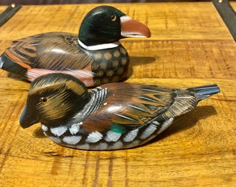 May include: Two hand-painted wooden duck ornaments. The ducks feature detailed feather patterns in brown, black, and white. One duck has a green head and a red beak, the other has a black head and a blue beak. They are displayed on a wooden surface.