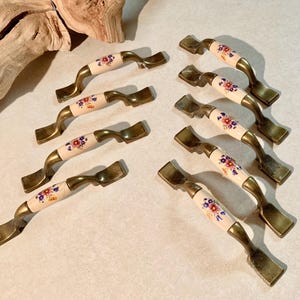 May include: A collection of eight vintage cabinet pulls. Each pull features a cream-colored ceramic center with a floral design in red, blue, and yellow. The handles are a brass color, and the pulls are arranged on a light-colored surface.