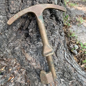 May include: A vintage bronze-colored ice axe with a T-shaped head and a curved pick. The tool is resting against a tree trunk, showcasing its aged patina and design. The axe is a classic mountaineering tool.