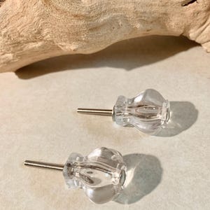 May include: Two clear glass cabinet knobs with a flower-like design. Each knob has a silver-colored screw for installation. The knobs are displayed on a light-colored surface, with a piece of driftwood in the background.