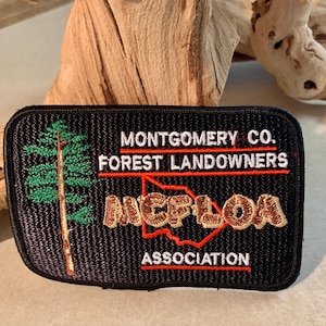 May include: Black rectangular patch with the text "MONTGOMERY CO. FOREST LANDOWNERS" in white and red. The patch features a green tree and the letters "NCFLOA" in tan, with a red outline of a state. The word "ASSOCIATION" is in white.