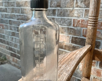 May include: A clear glass bottle with a black cap. The bottle has measurement markings on the side. The bottle is sitting on a wooden surface in front of a brick wall.
