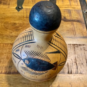 May include: A handcrafted gourd vase with a dark blue top and a beige body. The body features intricate carved designs, including a fish motif and geometric patterns. The vase is displayed on a wooden surface.