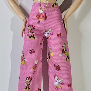 Op de afbeelding: Pop met een zwarte crop top, een gouden ketting en een roze broek met wijde pijpen. De broek heeft een herhaald patroon van Minnie Mouse, Daisy Duck en andere Disney-thema's. De pop draagt zwarte schoenen.