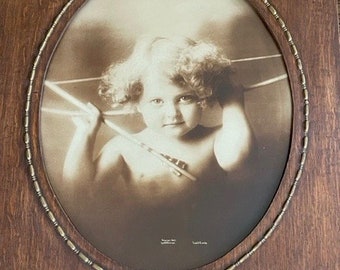 Puede incluir: Retrato en tonos sepia de un niño con cabello rizado, sosteniendo flechas, enmarcado en un óvalo con un marco decorativo de metal dorado. El marco está dentro de un marco rectangular de madera oscura, creando una estética vintage.