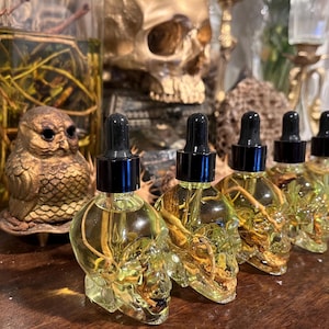 May include: A collection of skull-shaped glass bottles with black droppers, filled with a yellow liquid. The bottles are arranged in a row, with an owl figurine and a skull in the background. The bottles are likely for essential oils or perfumes.