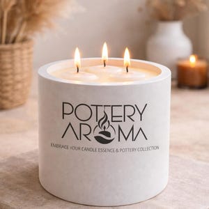 May include: A white cylindrical candle with three lit wicks. The candle has the text "POTTERY AROMA" and "EMBRACE YOUR CANDLE ESSENCE & POTTERY COLLECTION" printed on the front. The candle is on a stone surface.