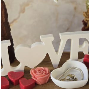 May include: A wooden tray displays a white "LOVE" sign, red heart-shaped candles, a pink rose-shaped candle, and a white heart-shaped dish holding jewelry. White flowers and a brown decorative item are in the background.