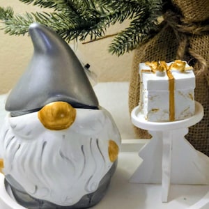 May include: A ceramic gnome figurine with a silver hat and white beard, accented with gold details. A small white gift box with gold ribbon sits on a decorative stand. Festive holiday decor.
