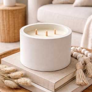 May include: A large, white, cylindrical candle with three wooden wicks. The candle sits on a stack of books and a wooden tray, alongside decorative beads and dried plants. The overall aesthetic is neutral and minimalist.