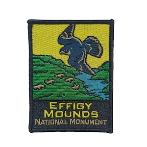 Op de afbeelding: Een geborduurde patch in zwart en geel met een blauwe vogel die over een groen landschap met een blauwe rivier vliegt. De tekst "Effigy Mounds National Monument" staat op de patch.