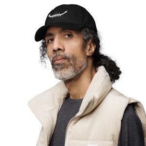 May include: A man wearing a black baseball cap with a white design on the front. He is wearing a beige corduroy waistcoat over a grey shirt.